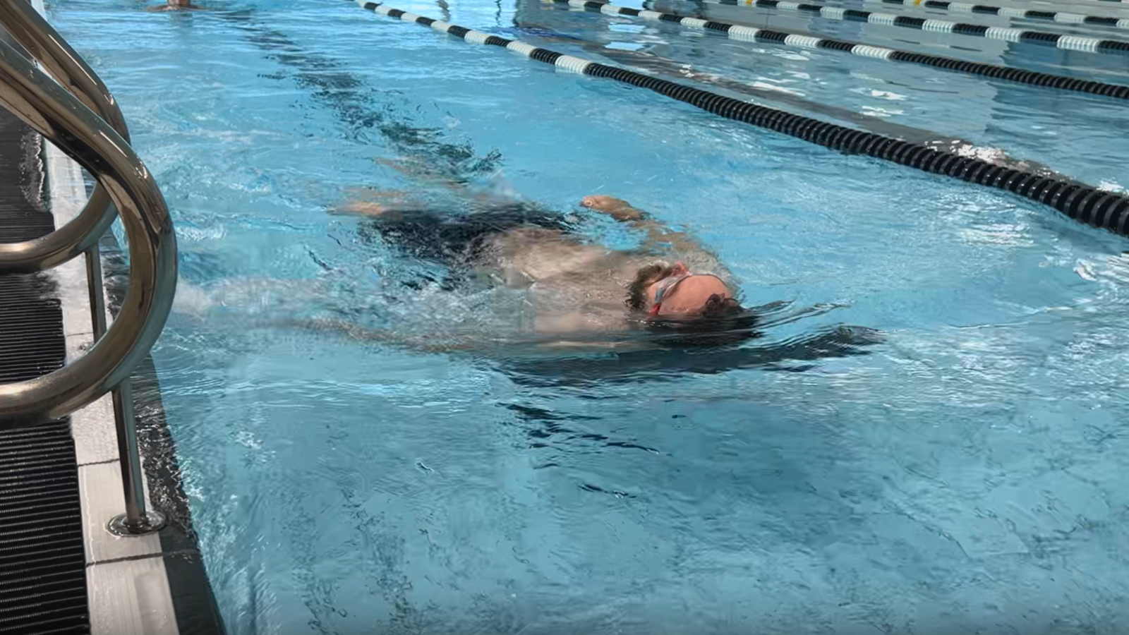 A man wearing black swimming trunks and red swimming goggles, swimming lengths of backstroke in an indoor swimming pool.