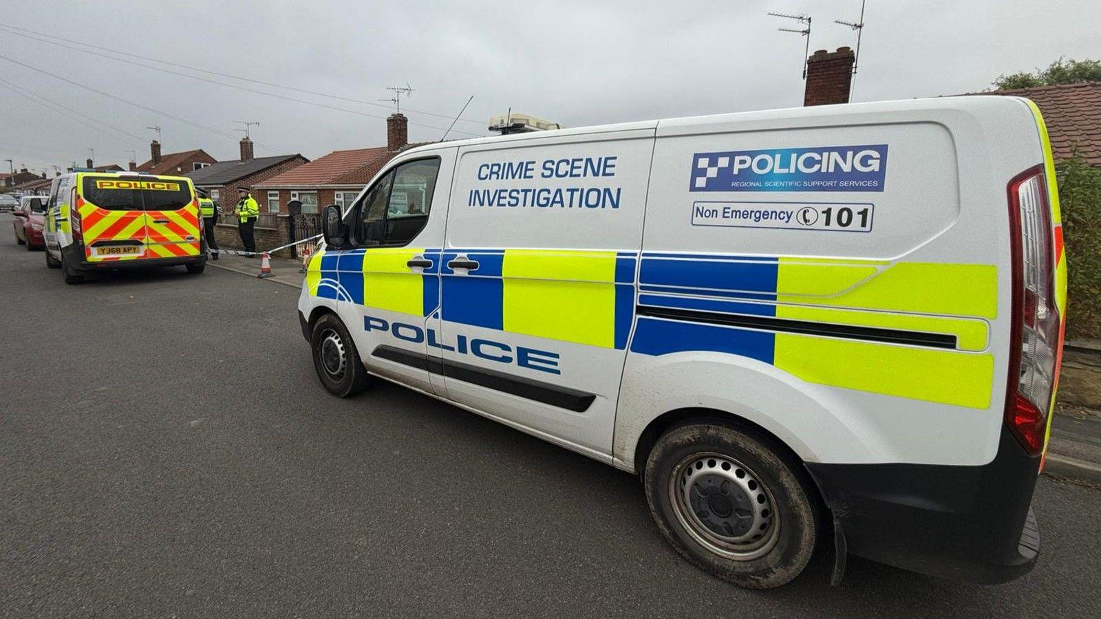 A white police van is parked in front of a house which has been cordoned off with blue and white police tape. Two officers can be seen in the background standing next to a second police vehicle.