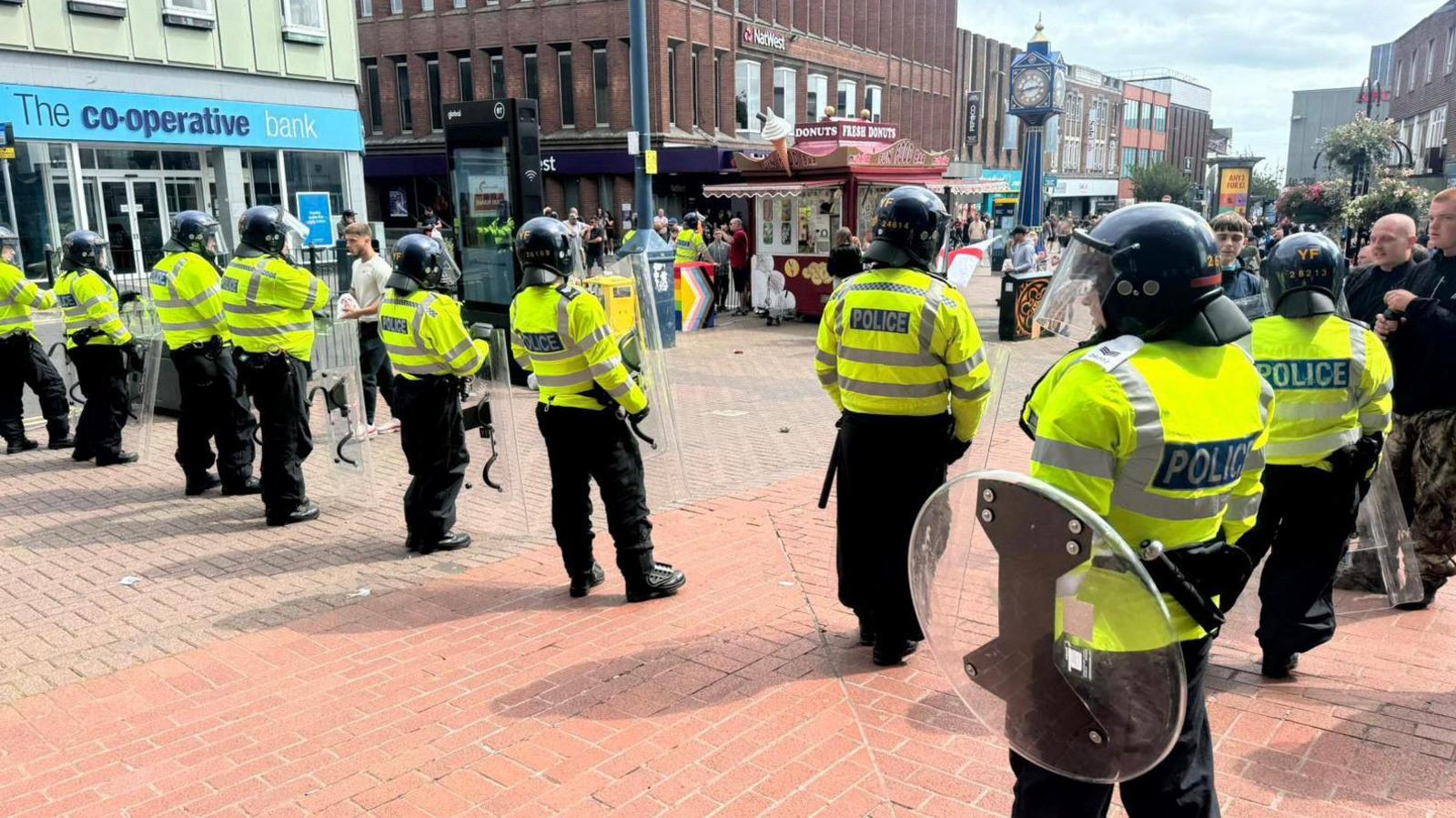 Riot police in Hanley in August. The officers are wearing green uniforms with "police" written on them and carry shields. They are in Hanley town centre with a clock tower in front of them and a parade of shops and businesses behind them, including the Co-operative Bank. They are standing on pink paving stones. 