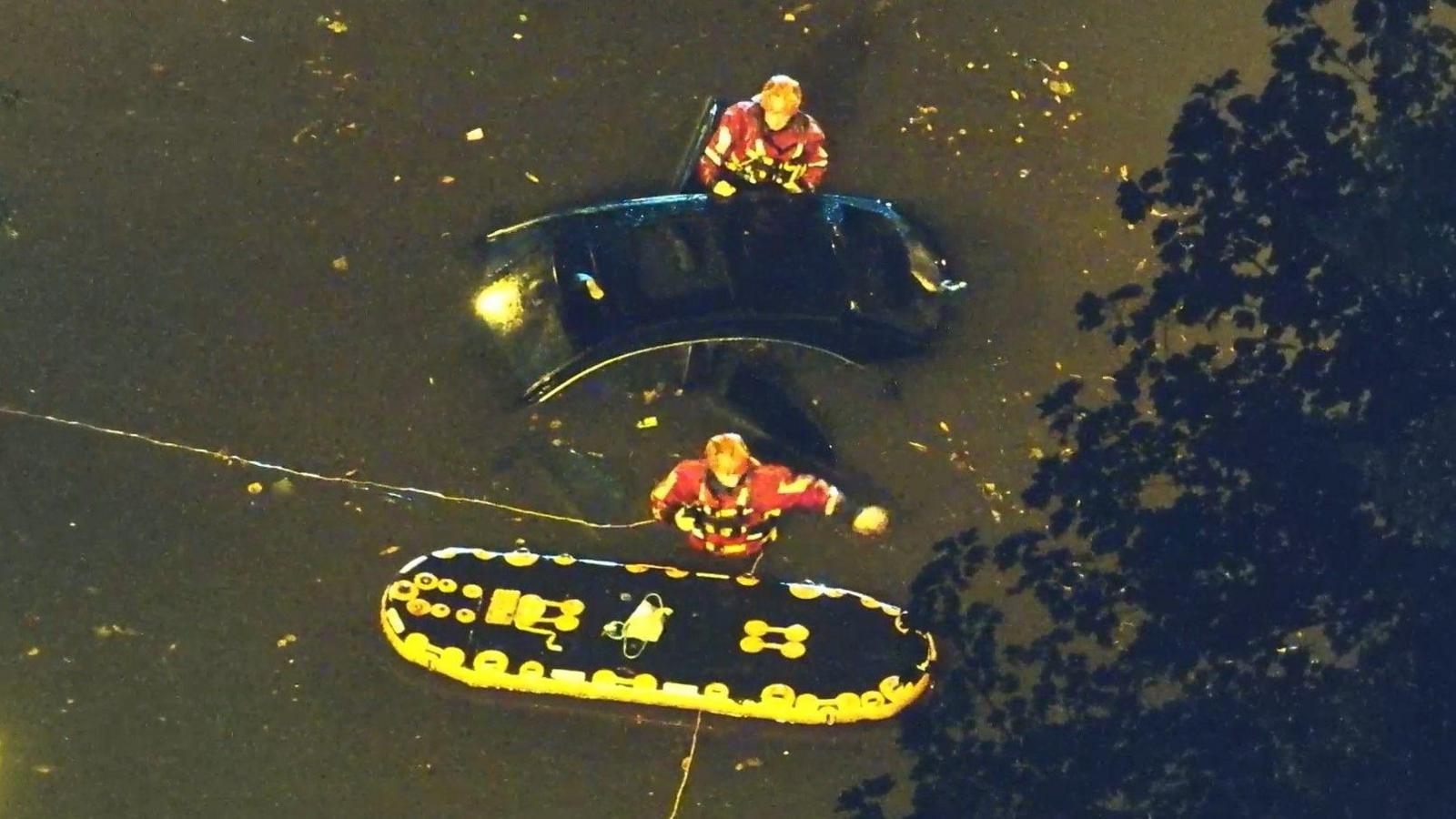 Drone footage showing two firefighters in red waterproof jackets and helmets waist deep in water next to a black car and a black and yellow inflatable raft.