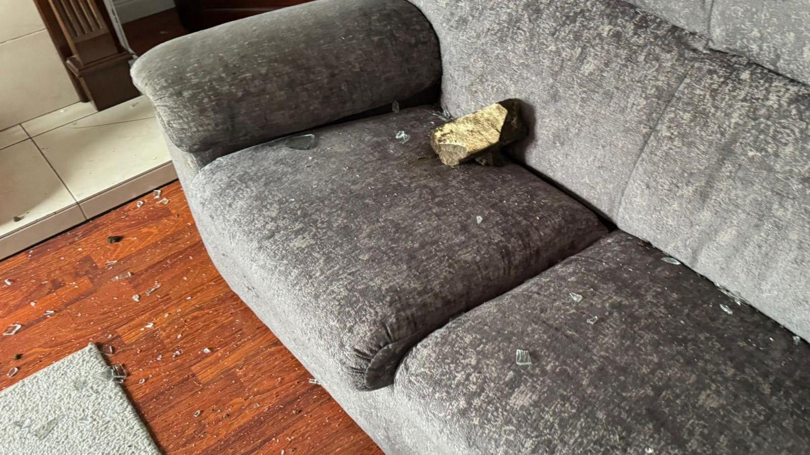 Grey sofa with a brick on it and glass and brick shattered on sofa and floor 