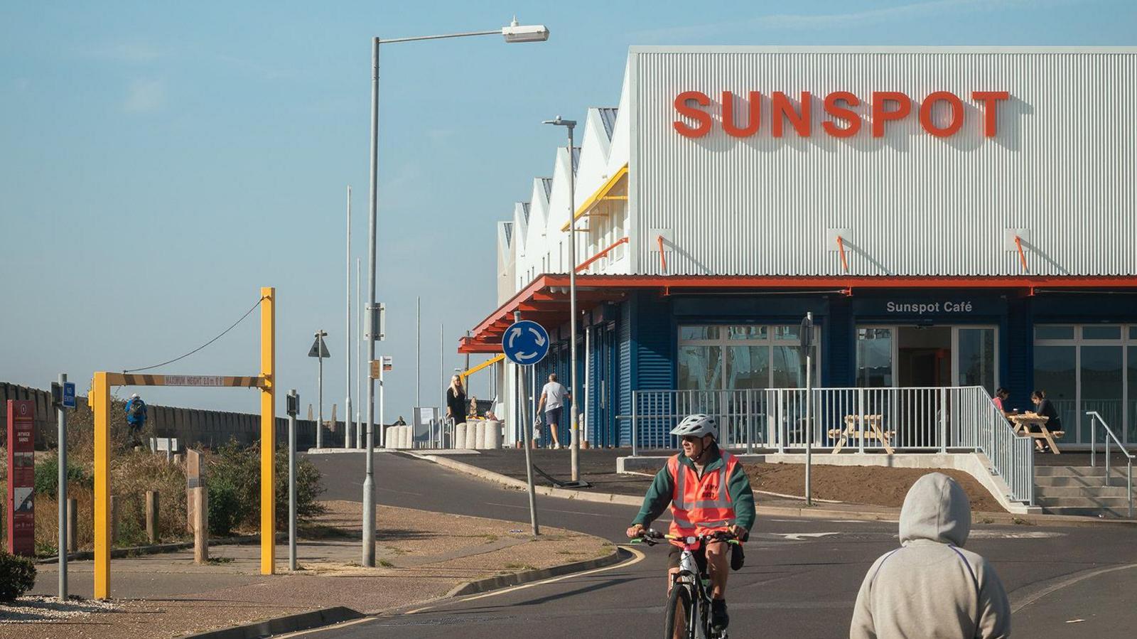 A two-storey building with a white upper floor with pointed roofs and the word SUNSPOT in red lettering on the front. There is a red dividing roof between it and the lower floor, which is dark grey in colour and includes tall windows and doors. A person in a white cycle helmet has just ridden past it.
