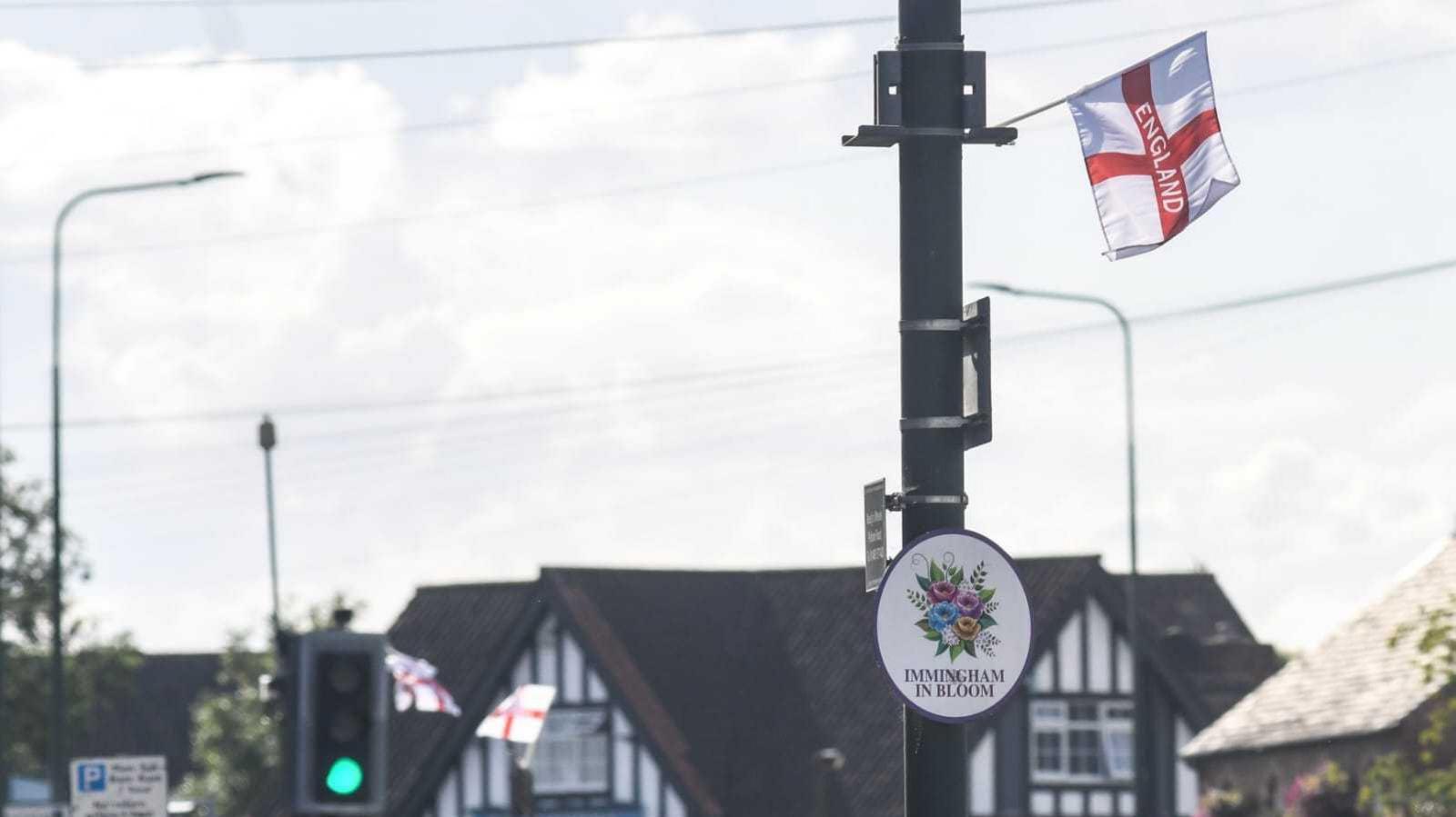 A small England flag with a white square, red cross and letters ENGLAND flying off a lamppost, which has a sign saying 'Immingham in Bloom' on it with flowers.