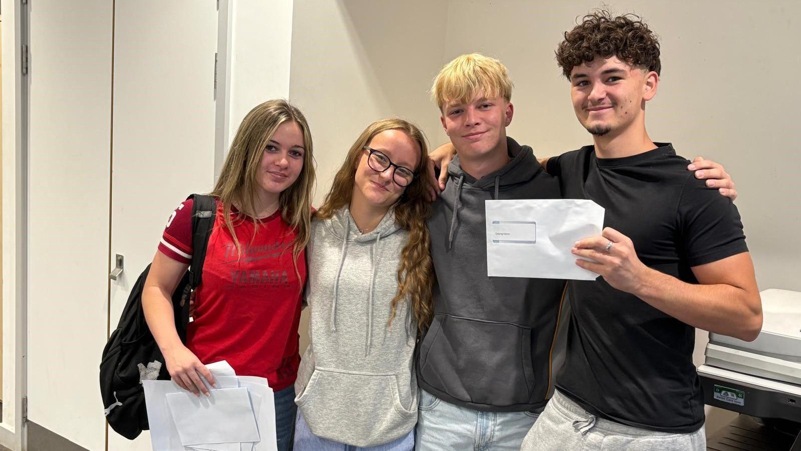 Four young people, two girls and two boys, link arms and hold up their GCSE results for the camera