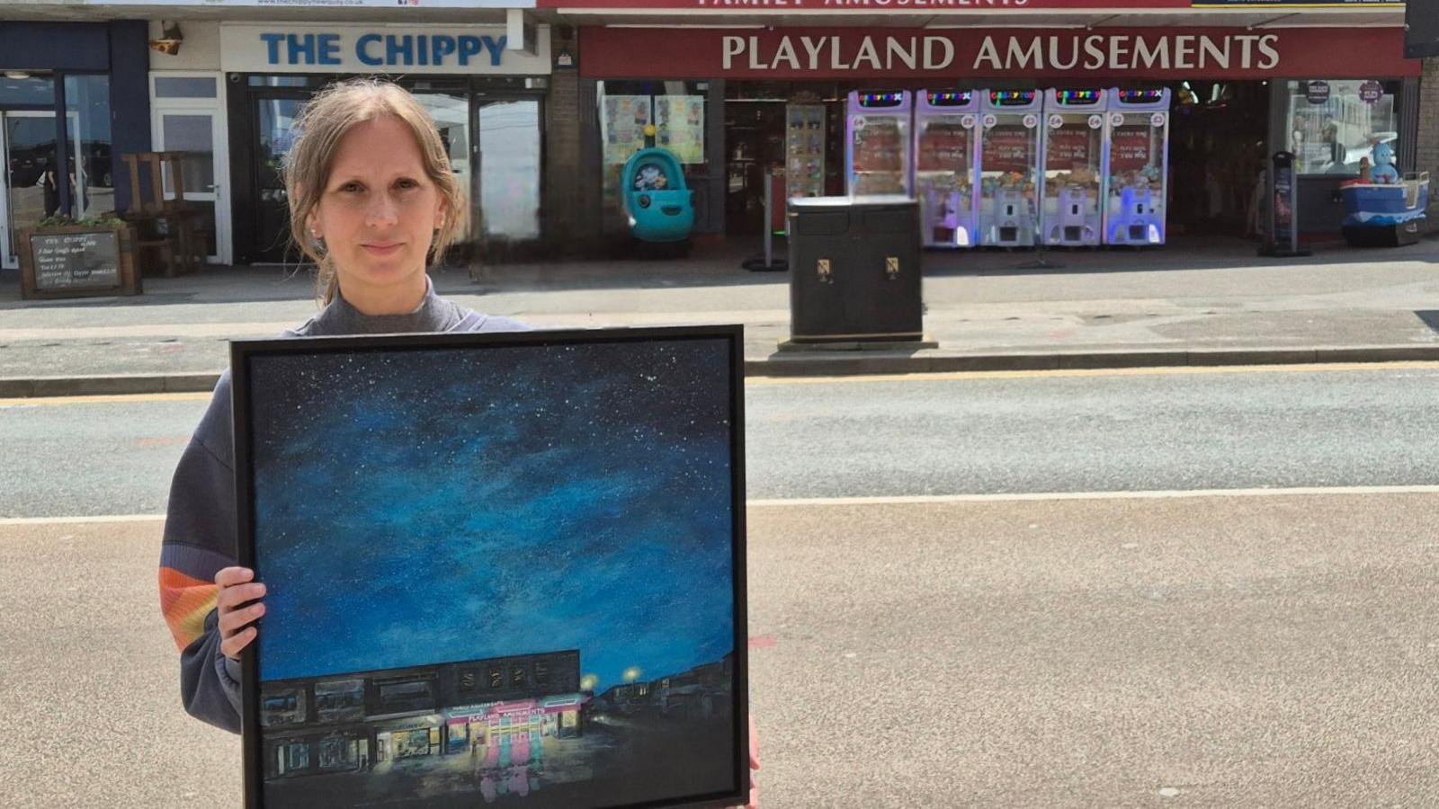 A woman stands outside an amusement arcade, holding a painting of the an amusement arcade at night, with a bright blue starry sky above