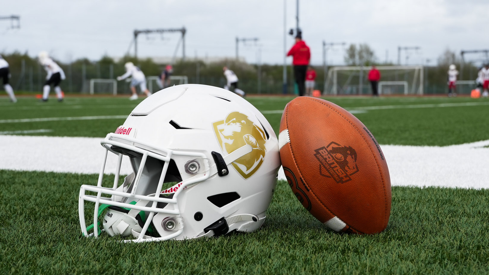 An American football and helmet placed next to each other on a pitch. People are playing American football in the background.