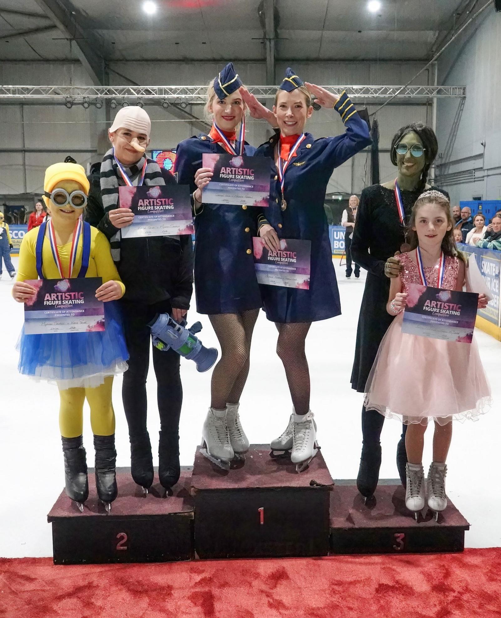 Six ice skaters stand on a podium. On the left, in the second place position is a child dressed in yellow as a Minion and an older child to her right wears a Gru costume, from the film Despicable Me. In the middle, in the first place position, are two women in air hostess uniforms. On the right, in the third place position, are a woman and child dressed as Elphaba and Glinda from Wicked.