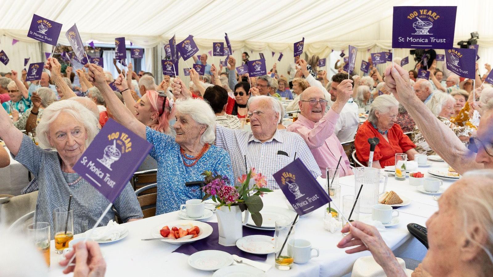 A large group of older people seated at long tables wave small purple flags as they celebrate the 100th anniversary of the St Monica Trust in Bristol. They are in a large marquee with a white roof and walls