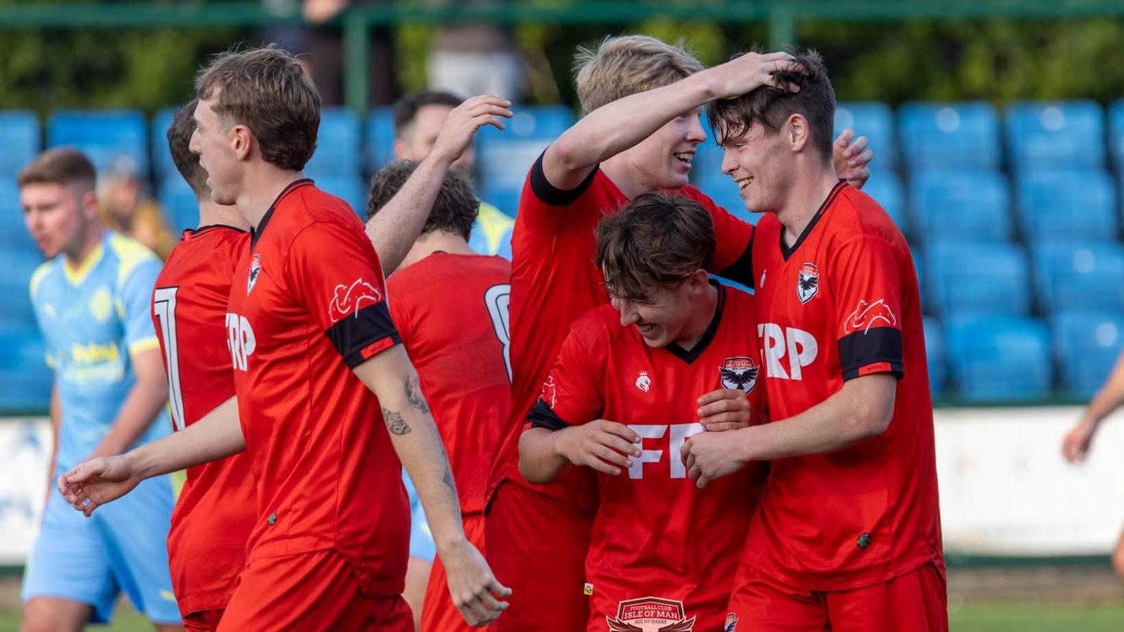 The Manx team wear a red strip and celebrate together after a goal. One man pats another team-member's head.