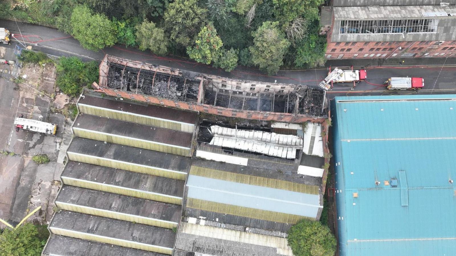 A drone photo of a warehouse building following a fire which burned out its roof. A large blue-roofed building is seen on the right, with fire engines parked in the street. Trees and other buildings line the road.