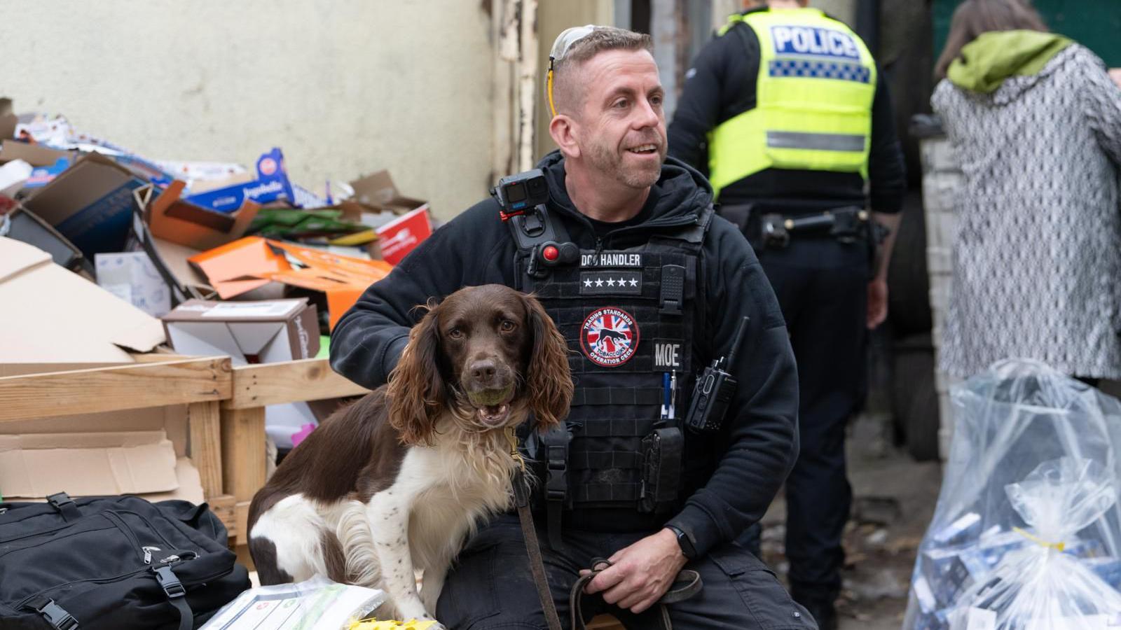 Stuart Philips has short grey hair and is wearing navy blue security-type clothing and a black padded vest. He is holding a lead with a brown and white cocker spaniel attached. They are sitting with a pile of cardboard boxes behind them. A police officer and another person wearing a grey coat are nearby.
