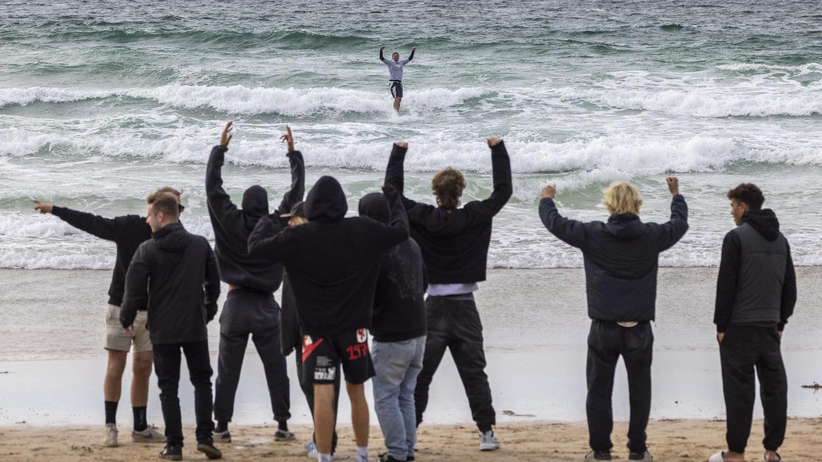 Several highly-animated people on a beach look out at a surfer. Most of them are waving their arms. The surfer has his arms up in celebration.