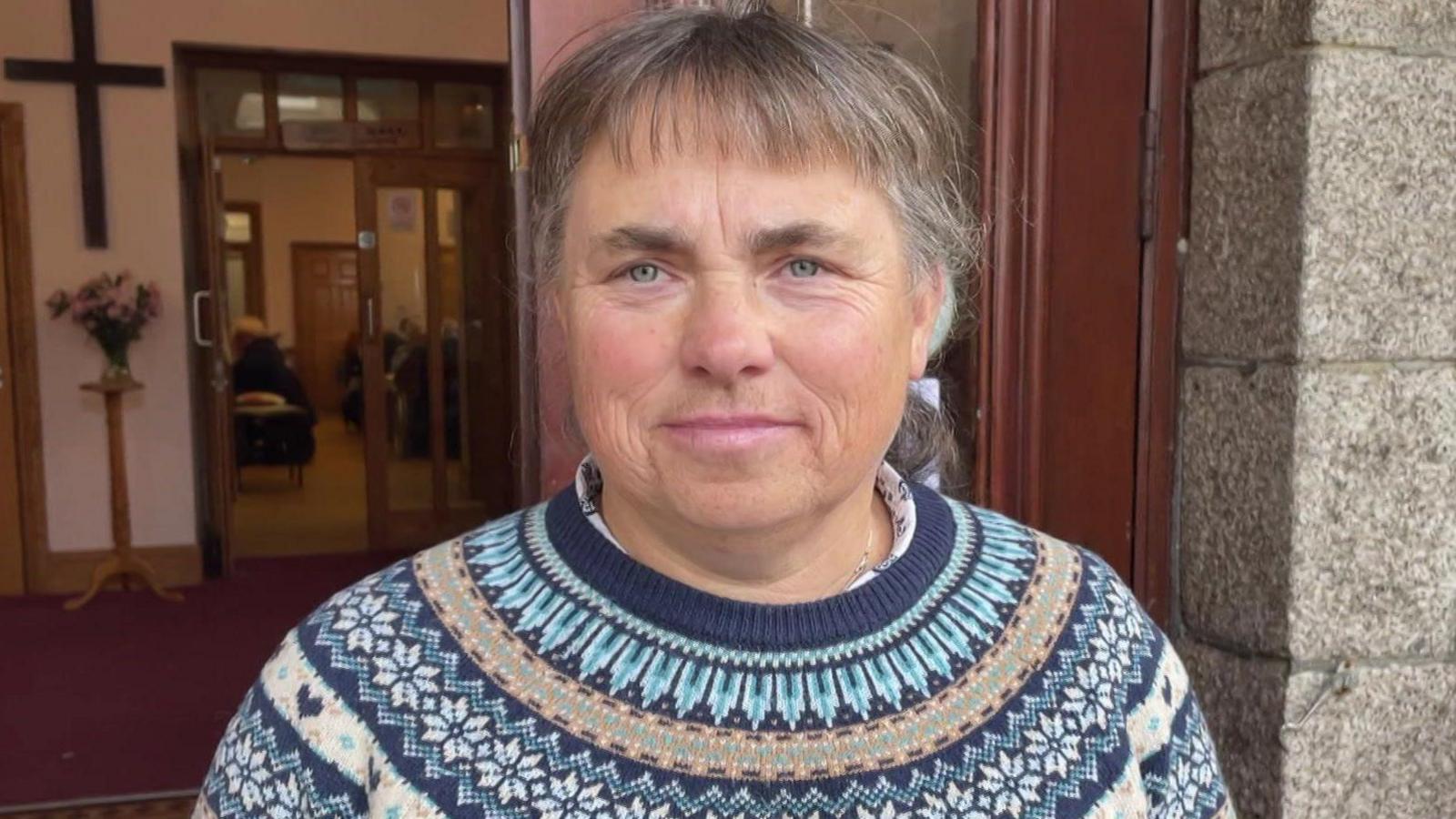 A woman wearing a navy blue jumper with a Scandinavian print on it of light blue, white and brown patterns. She is standing at the doorway of a church building.