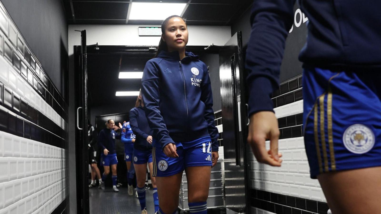 Asmita Ale camina antes del saque inicial de un partido de fútbol. Lleva la equipación del Leicester City y una chaqueta azul con cremallera encima.
