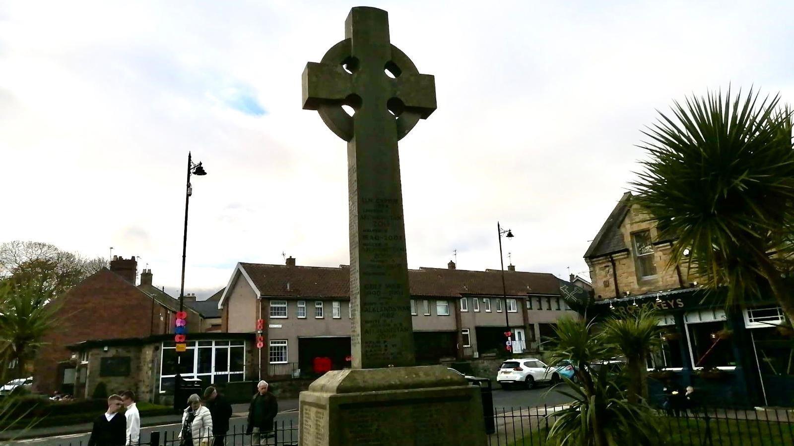 A large war memorial is in the centre, with railings around it. Behind is a row of housing.  