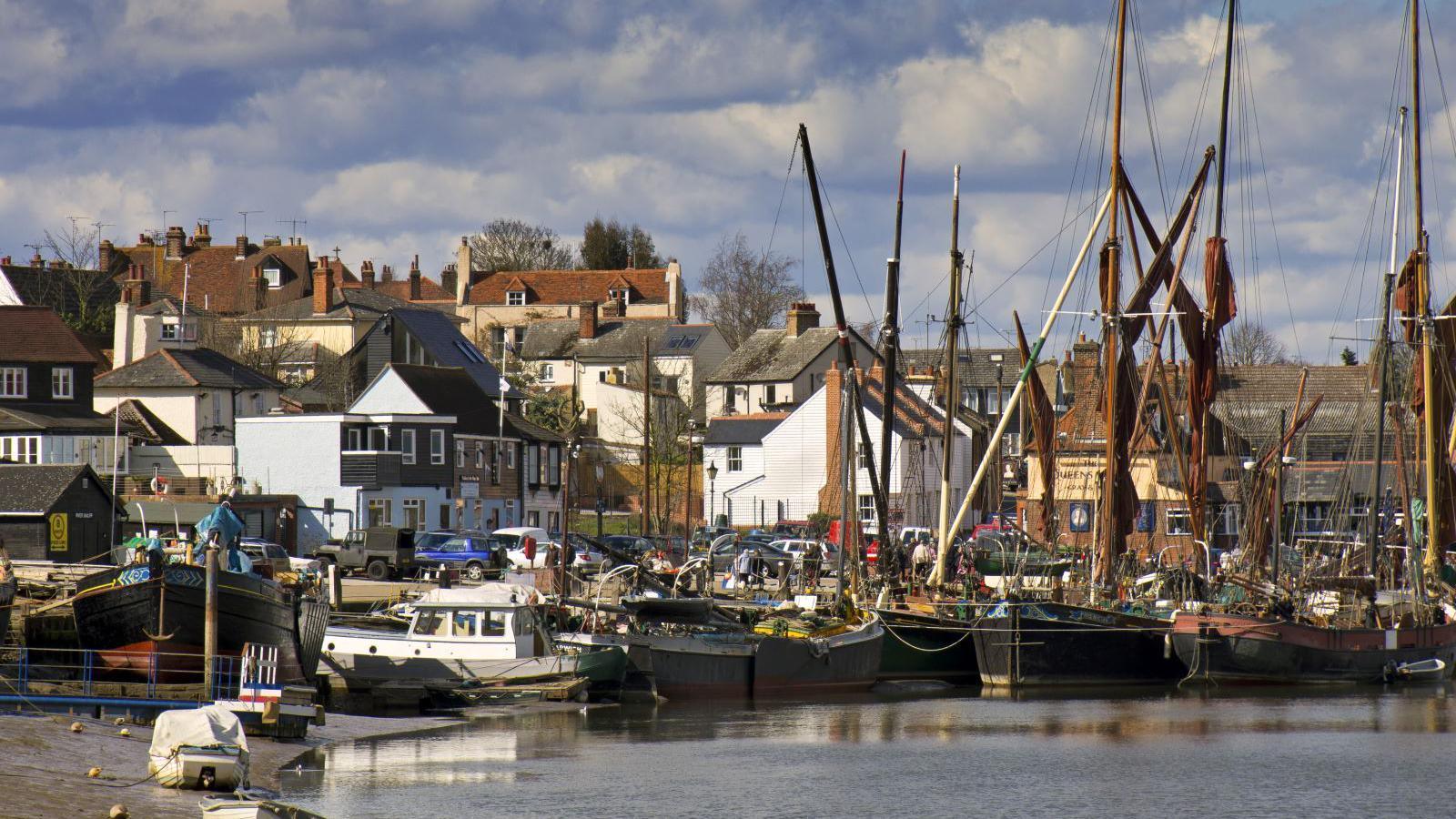 An array of ships docked in Maldon's quay. The picture has been taken at low tide on a bright but cloudy day. Houses and venues line part of the water front.