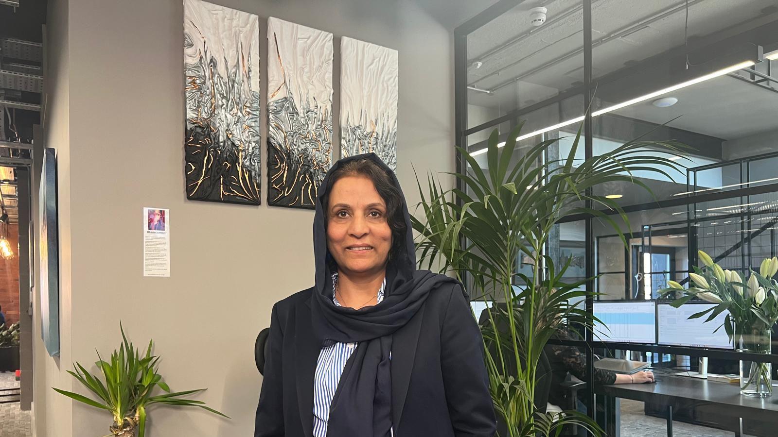 A woman in a navy blue blazer, navy blue headscarf and blue striped shirt is standing in front of three similar pieces of rectangular art in an open place office space. There are also tree shrubs on either side of her.