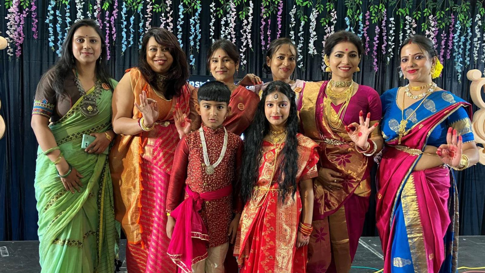 Six women and two children, wearing traditional Bengali outfits stand on a stage. Some have red paint on their hands and tips of their fingers