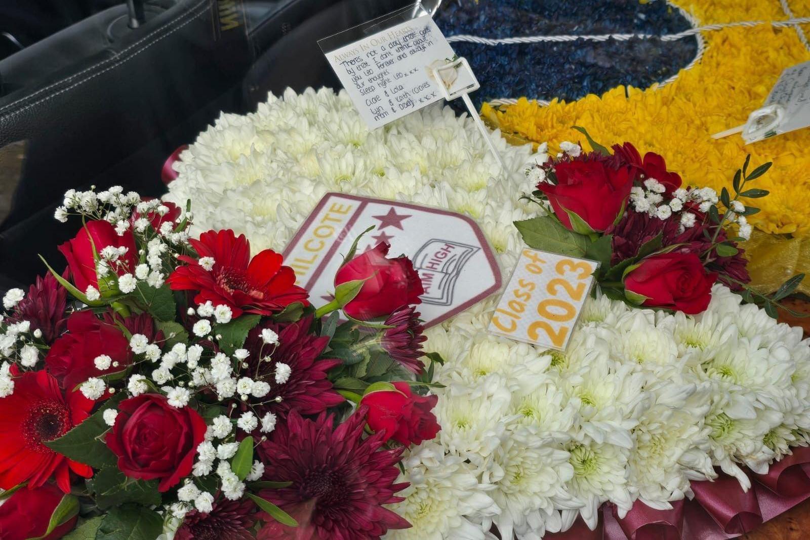 A close-up of floral wreaths. A large white wreath is in the centre with a school crest on top which says Class of 2023. Red flowers are placed to the left of the wreath with a few red flowers in the middle. Yellow and dark blue flowers are to the right. There is a white note attached to the central wreath with a hand-written note on.