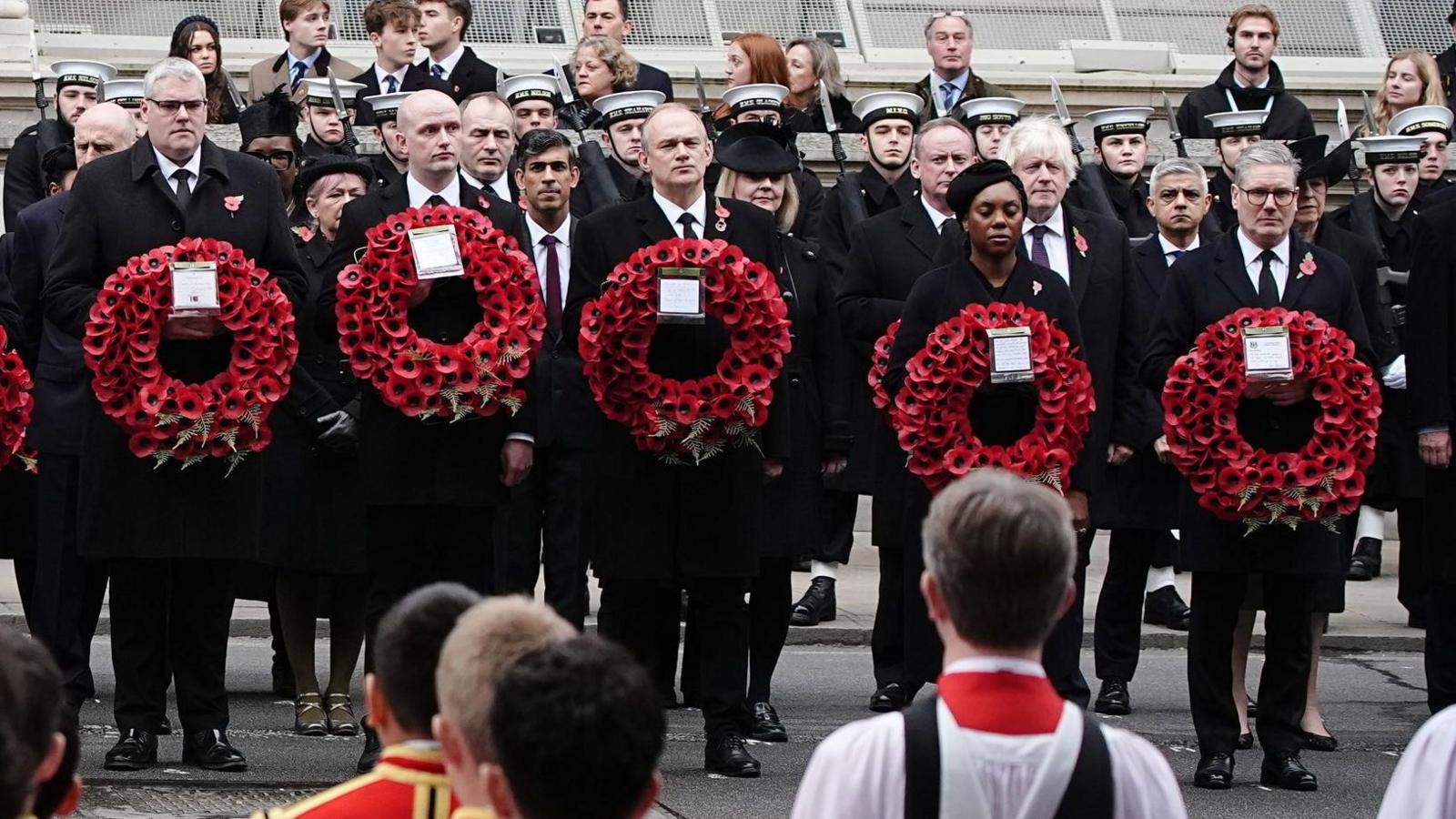 UK party leaders Gavin Robinson, Stephen Flynn, Ed Davy, Kemi Badenoch, and Sir Kier Stammer standing in a line holding poppy wreathes.