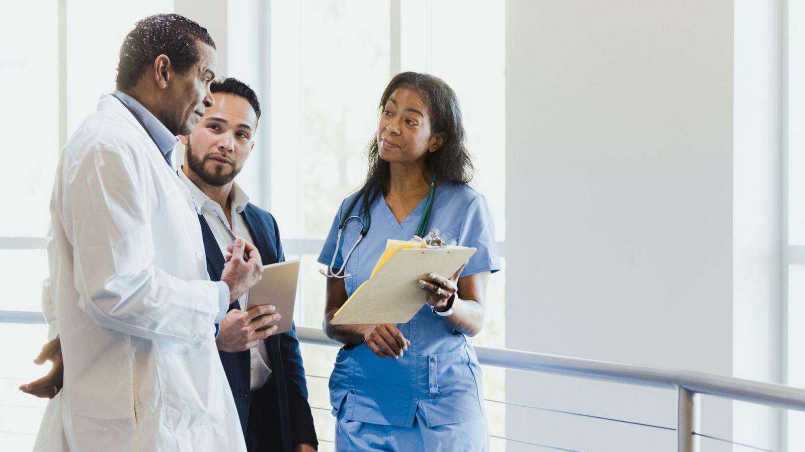 A senior doctor, wearing a white coat, is talking to a woman in blue uniform who is showing him some paperwork. They are standing in a hospital corridor painted white, next to some windows. A man in a suit stands between them.