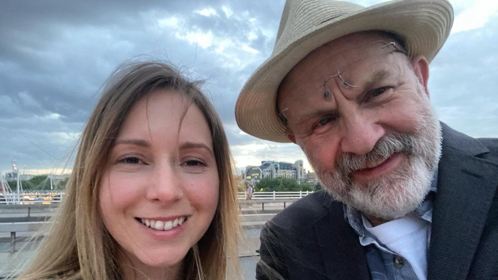 Carly Clarke has long blonde hair and Stephen Lovell-Davis is wearing a straw hat with his glasses on his forehead while wearing a black jacket and shirt.
A city skyline is visible behind them.