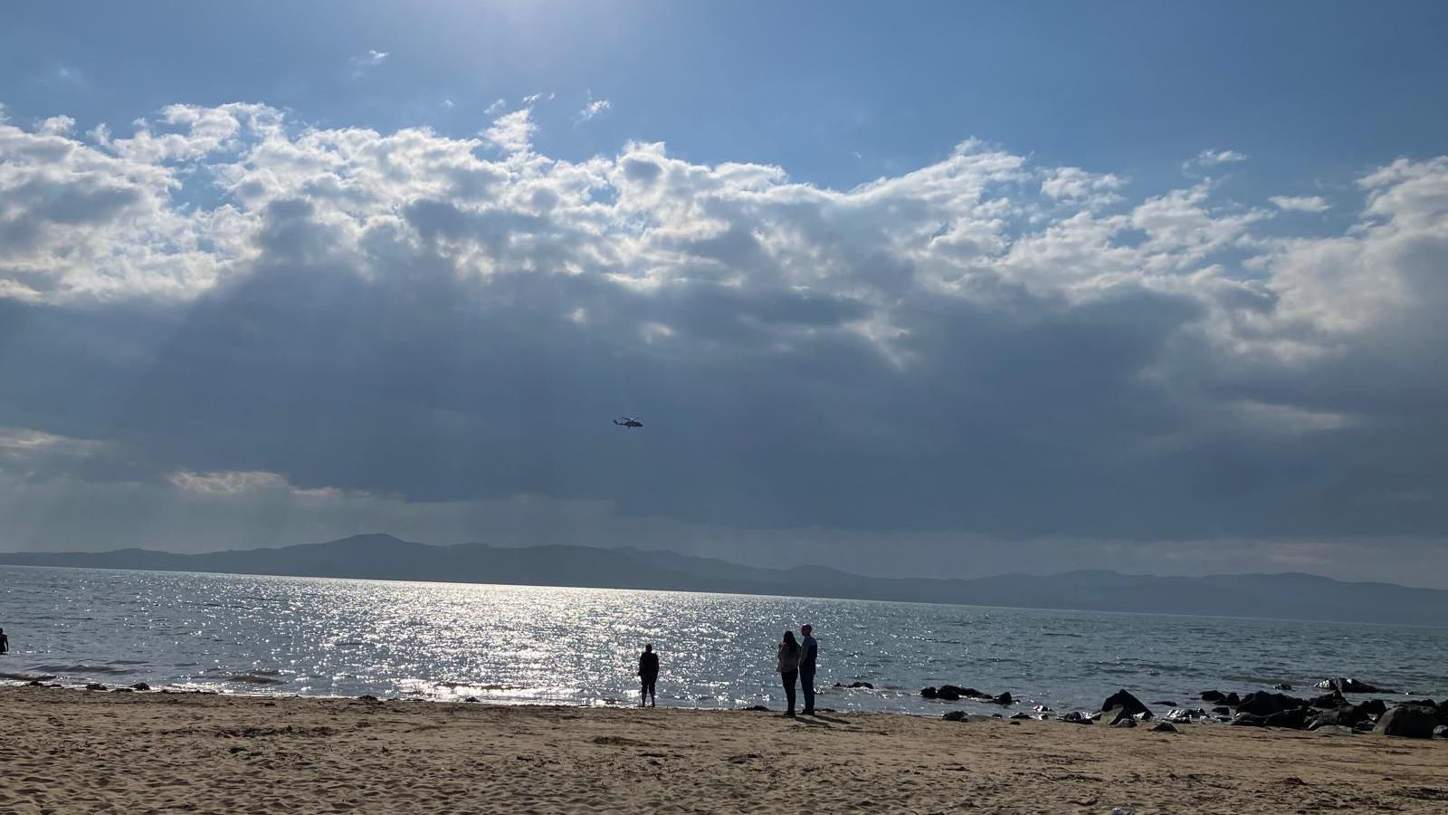 Three people standing on Buncrana beach in County Donegal are looking out to sea on a sunny day and in the sky a coast guard helicopter can be seen searching the water for swimmers who have been reported missing