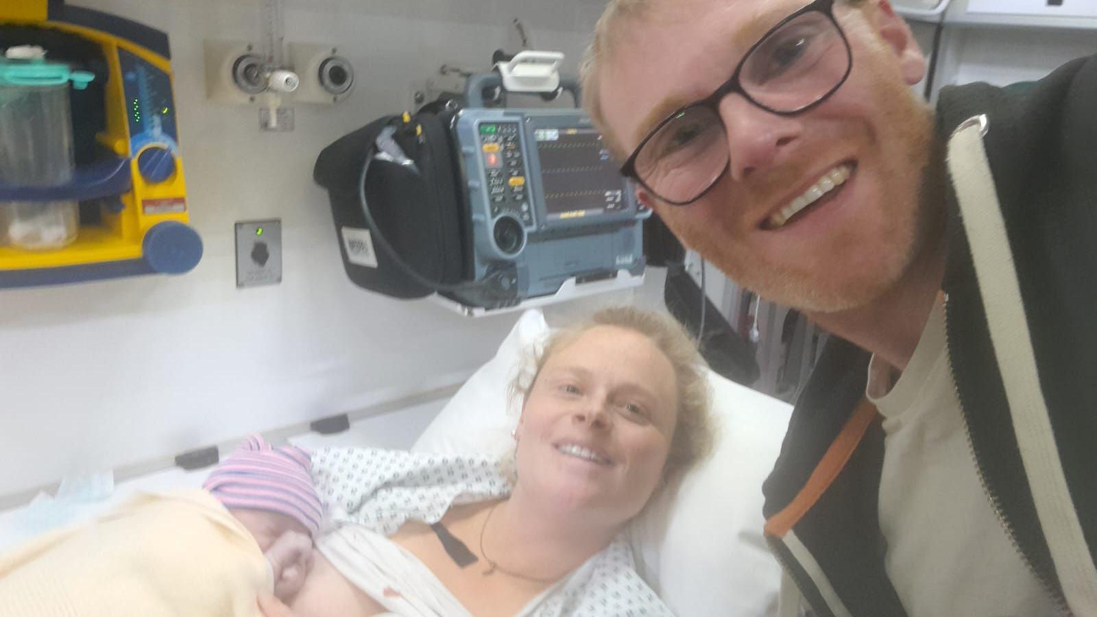 Ian, a man in his 30s with fair hair and wearing glasses, takes a selfie with Claire. She is lying in an ambulance bed as she smiles with their baby Tamzin wrapped up in her arms.