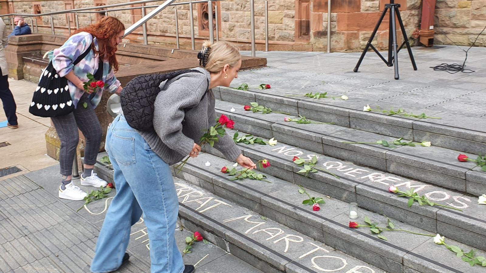 Two women lay flowers on the steps of Derry's Guildhall, there is Spanish language writing written on the steps