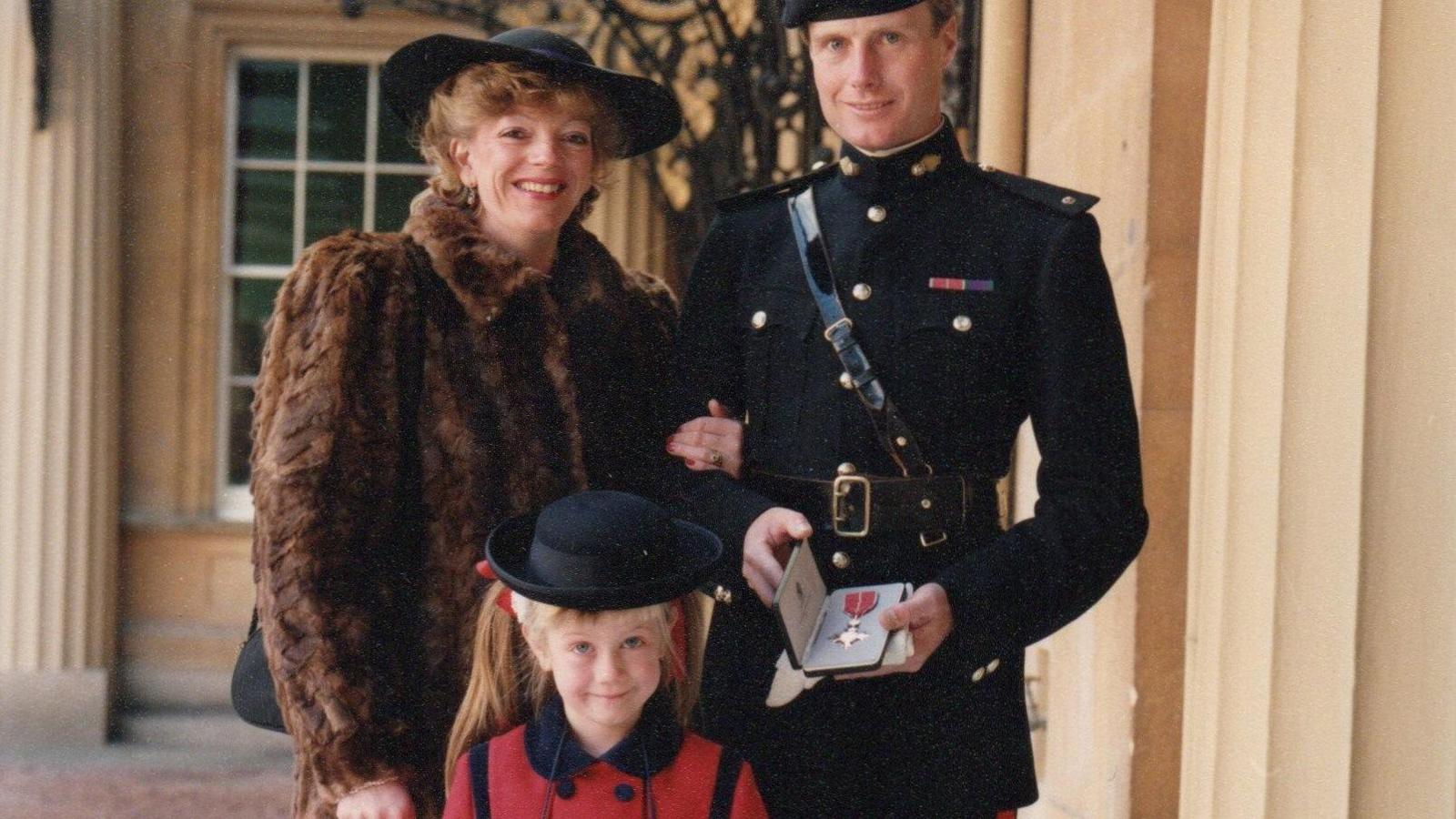 A man in formal black army uniform posed alongside a woman in a fur coat and hat while a little girl in a red and black coat and hat with long blonde ponytails stands in front of them. He is holding an MBE.