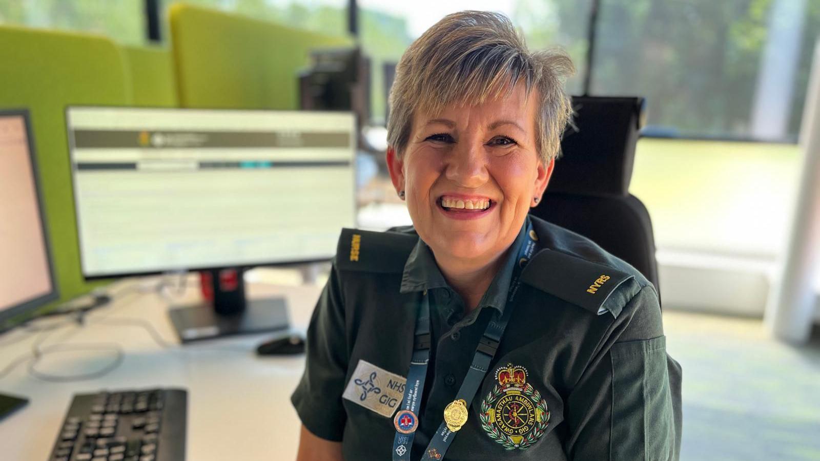 Amanda is wearing the green uniform of the ambulance service, with embroidered logos on it and a staff lanyard. She is sat at her desk with screens behind her. She is smiling at the camera.