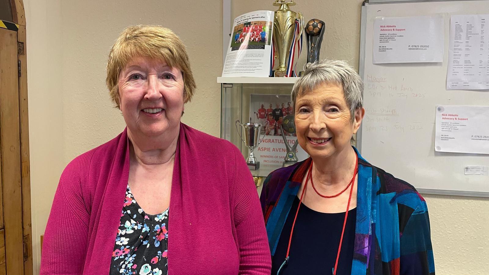 Two women, both with short hair, stand in front of a trophy cabinet. One woman is wearing a pink cardigan whilst the second, who is slightly shorter, is wearing a blue patterned cardigan. 