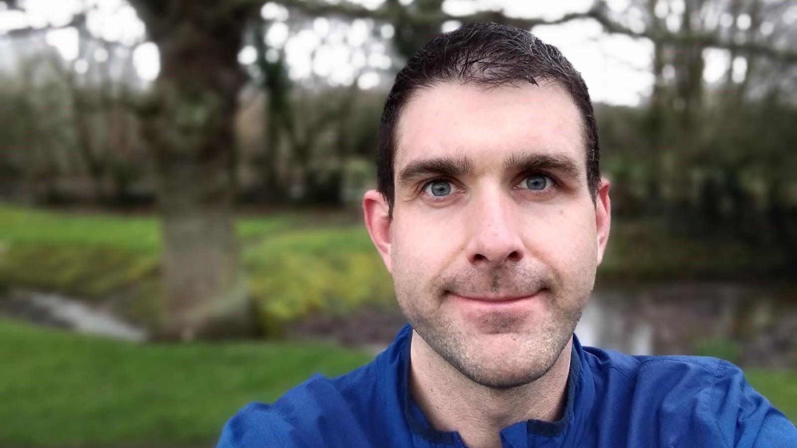 Selfie picture of Kieron in a park, wearing a blue zip-up top 