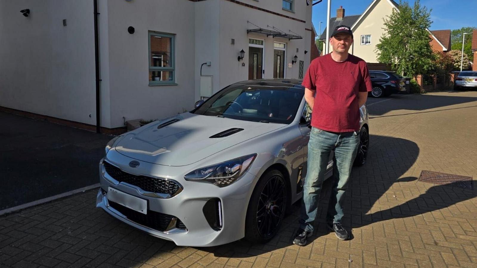 A man wearing jeans, a red t-shirt and a blue baseball cap is standing beside a grey car which is parked on a driveway outside a home