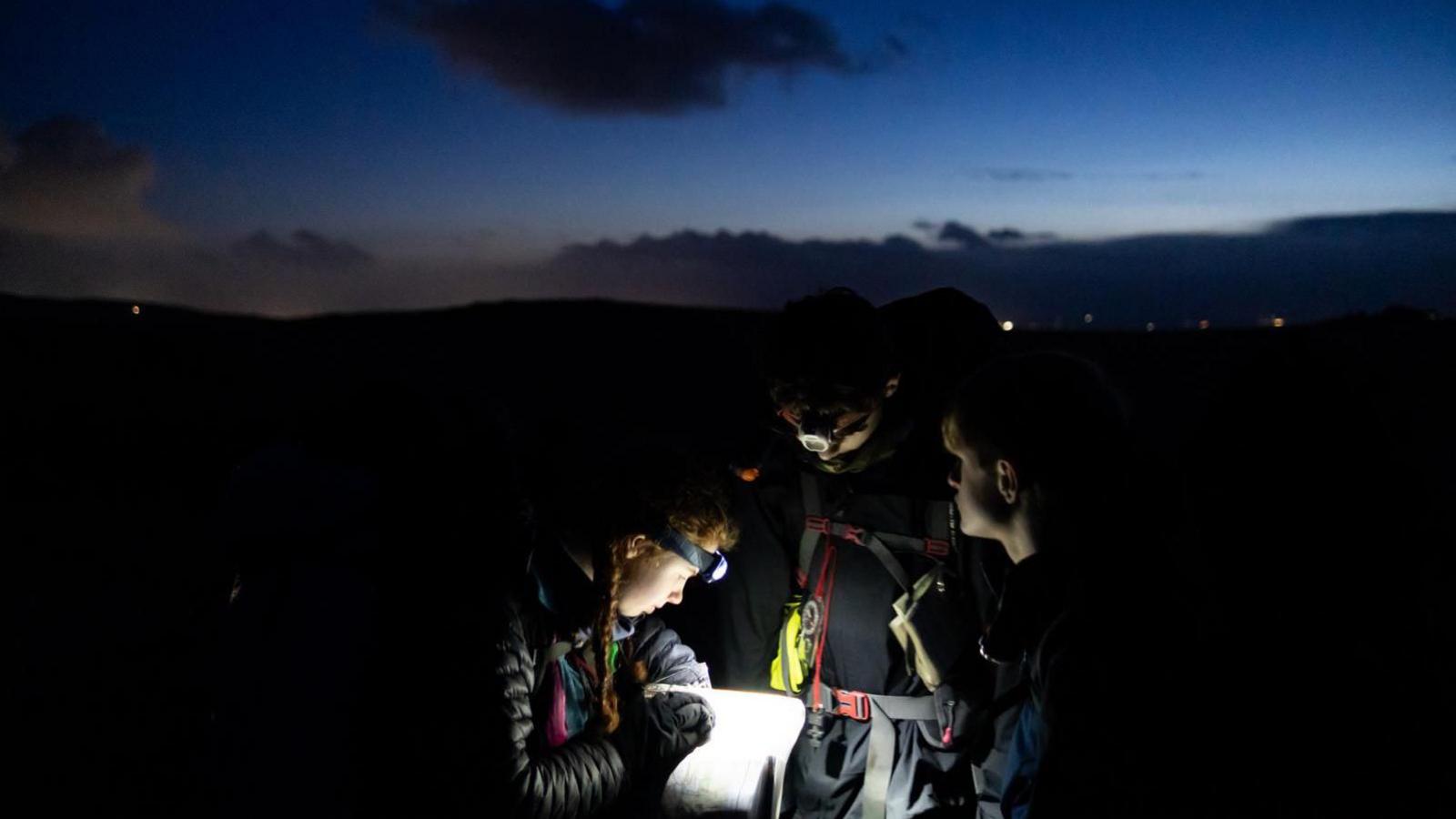 Three people stood in the dark with head torches lighting up a piece of paper.