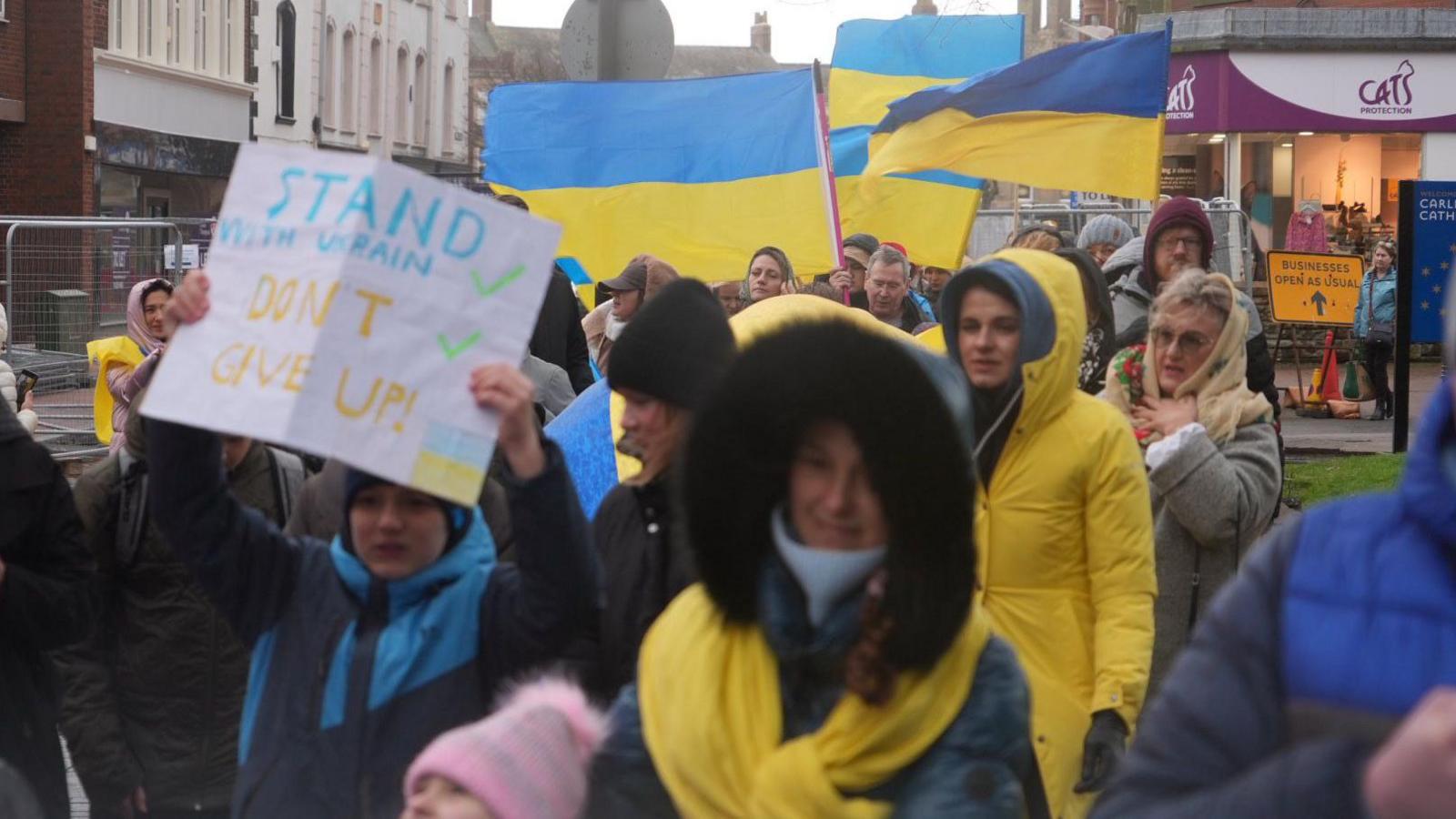 Dozens of people marching through Carlisle city centre. One is carrying a poster which reads "Stand with Ukraine. Don't give up". Several are wearing yellow or blue. A number are also carrying flags of Ukraine, which is a blue stripe on top of a yellow stripe.