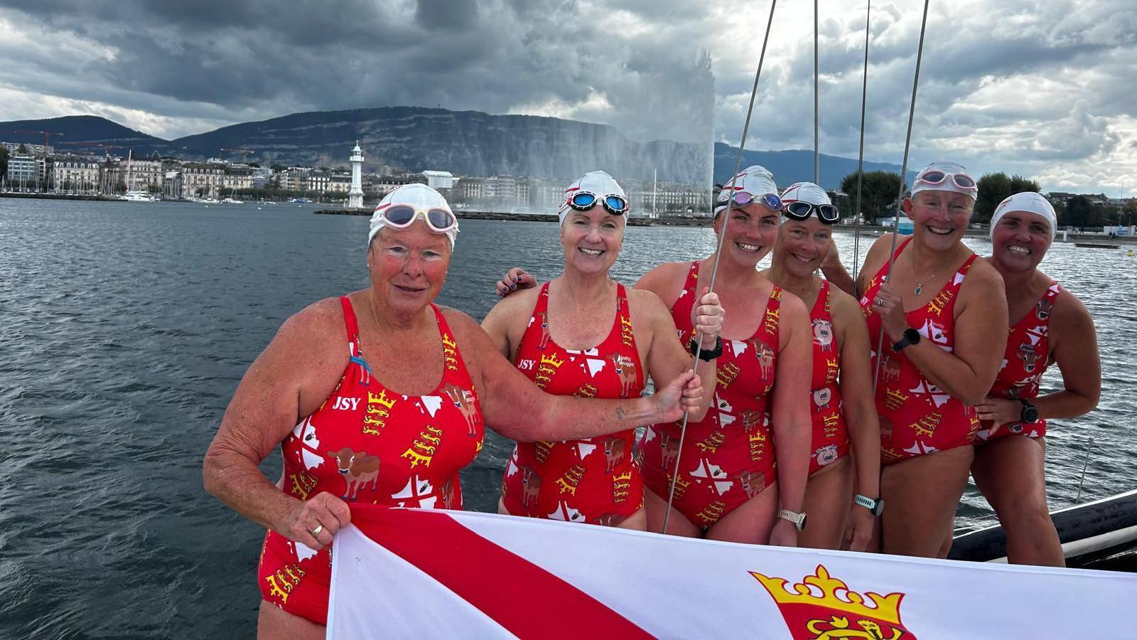 The six members of the swimming team smile at the camera in their swimwear in front of Lake Geneva. They're wearing red swimming costumes with Jersey logos, flags and cows on it. They're holding a Jersey flag between them.