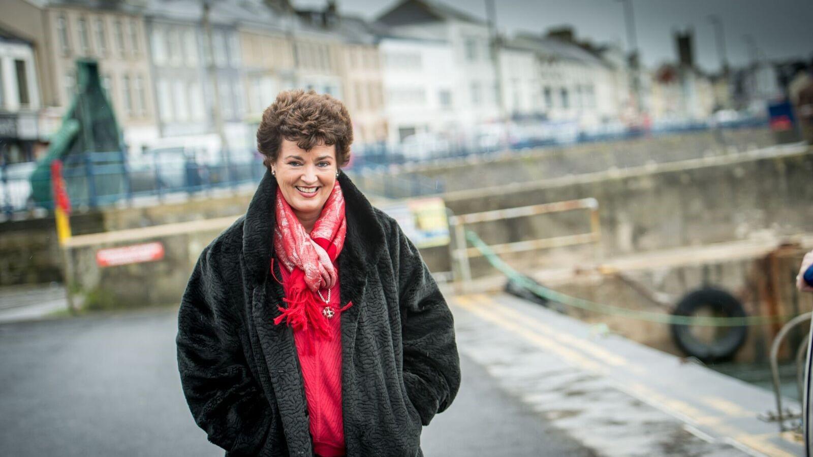 A woman with brown hair wearing a red jumper and a black jacket stands on a quay, smiling at the camera. Behind her is a row of houses. 