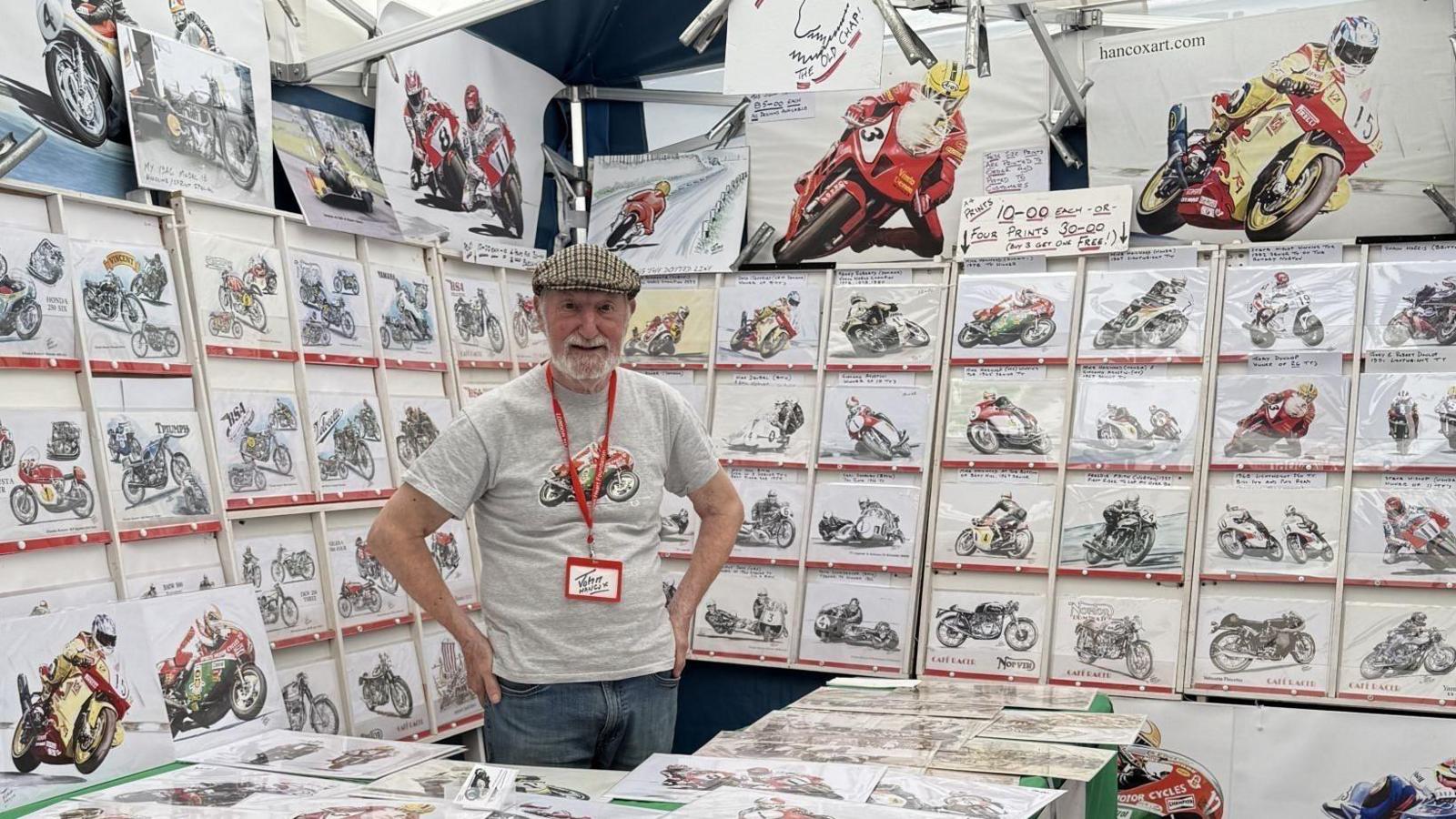 John Hancox, who is wearing a grey T-shirt with a drawing of a motorbike on it, surrounded by lots of examples of his work, which is also drawings of motorcycles.