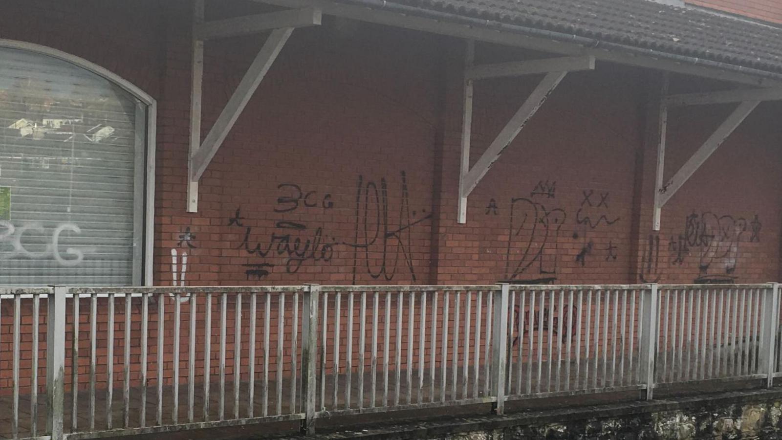 The image shows a red brick wall surrounded by a grubby white railing. Paint is peeling from white rafters. The walls have graffiti tags scrawled across them, in black. One tag appears to read "waylo", another says "3CG", while others are indecipherable. 