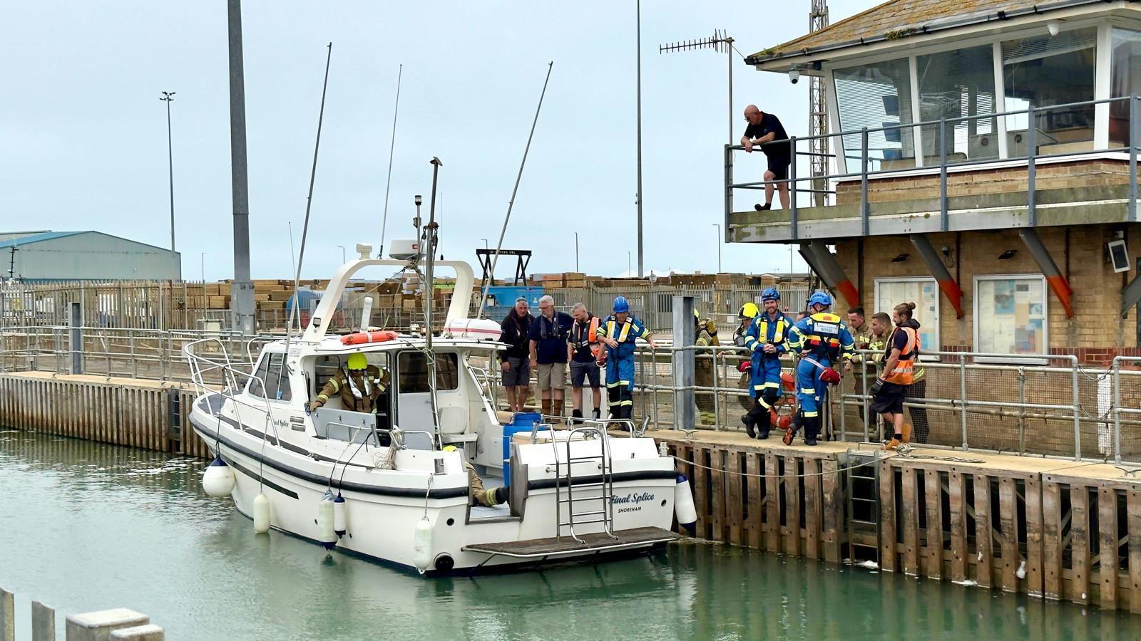 A white yacht sits in Shoreham Harbour, with emergency teams wearing blue and yellow standing on the boat and harbour, and two civilians standing on the yacht's deck.