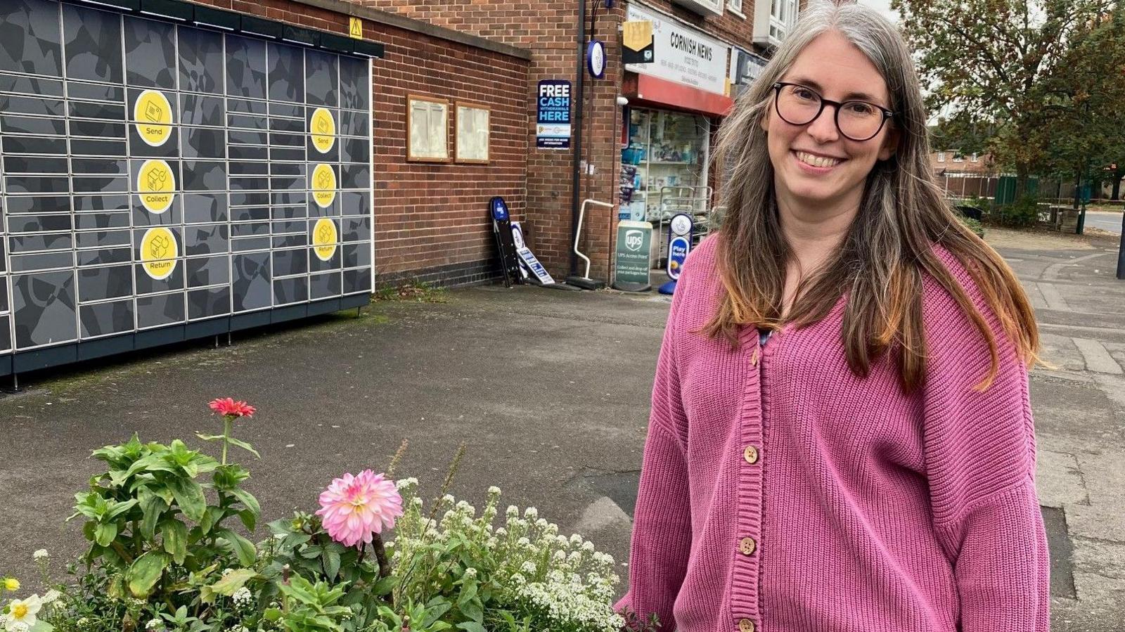 A woman stands in front of a row of shops next to a flowerbed full of plants and flowers. 