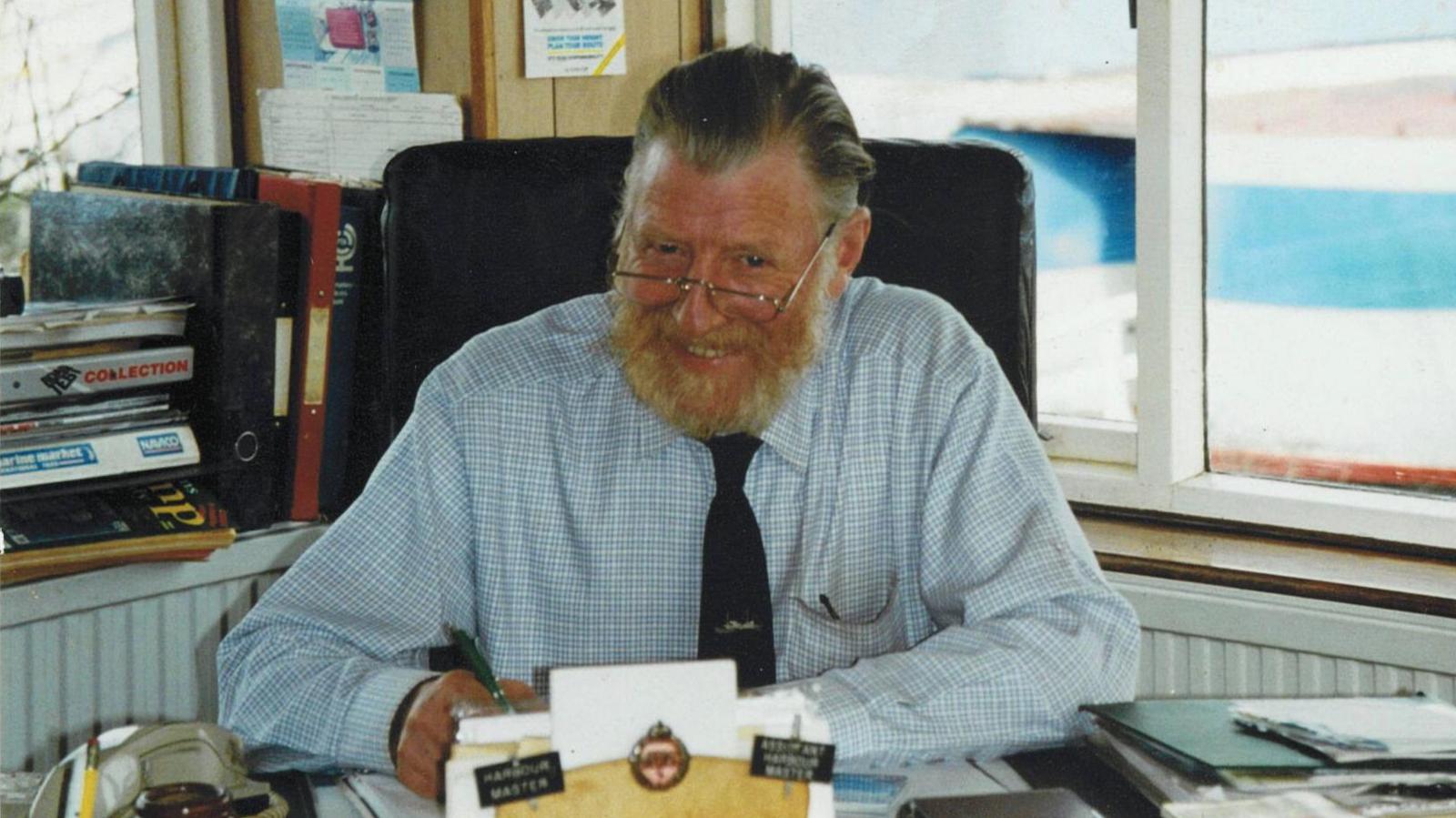 An photographer of Mark Ainsworth wearing a blue shirt and black tie, sat behind an office desk, smiling and glancing up at the camera while he works