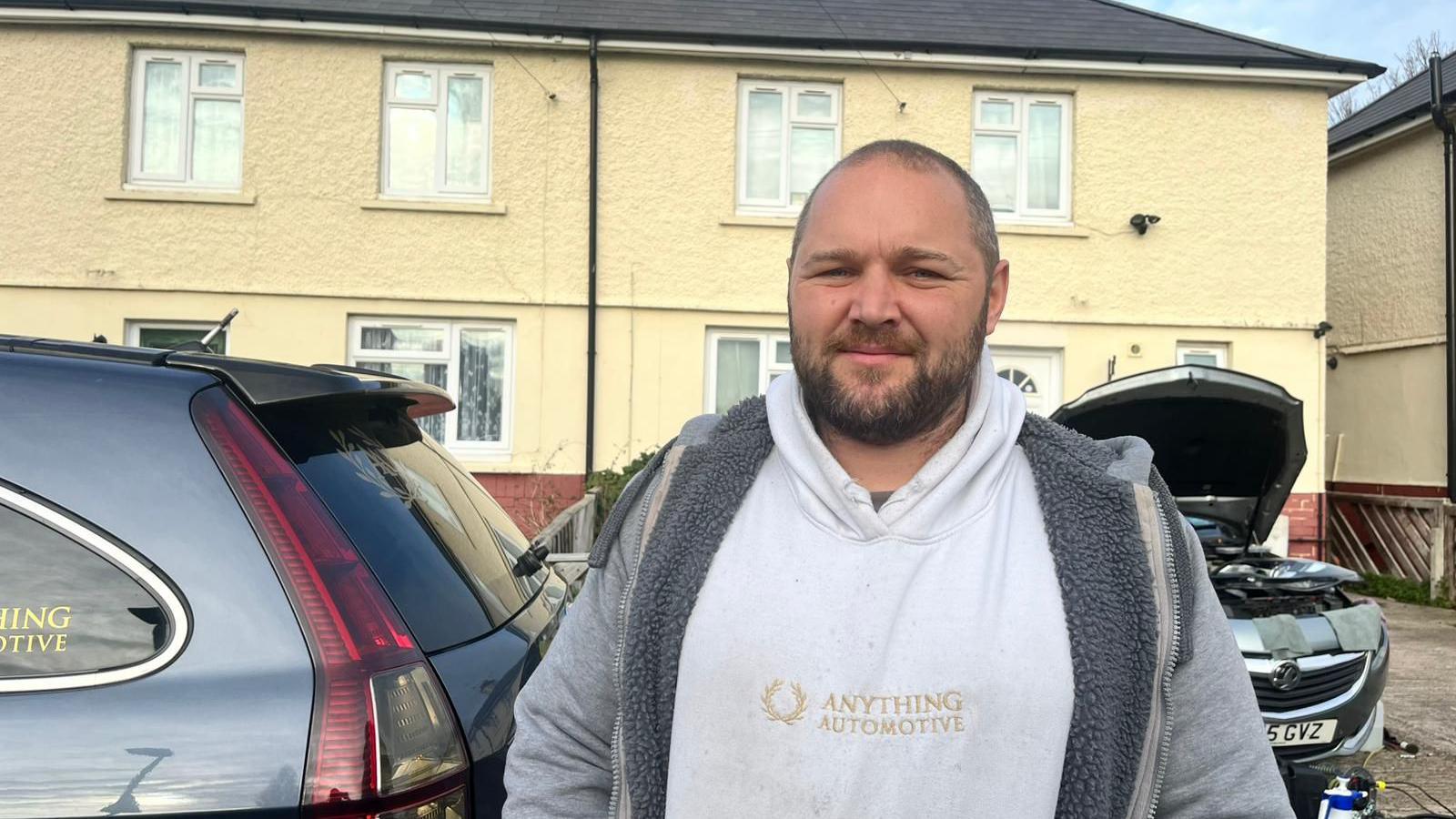 A middle-aged man wearing a white hoodie with a grey zip jacket on top. He has a serious expression on his face. He is stood in front of two silver parked cars, one with its bonnet up. Behind them are two attached houses.
