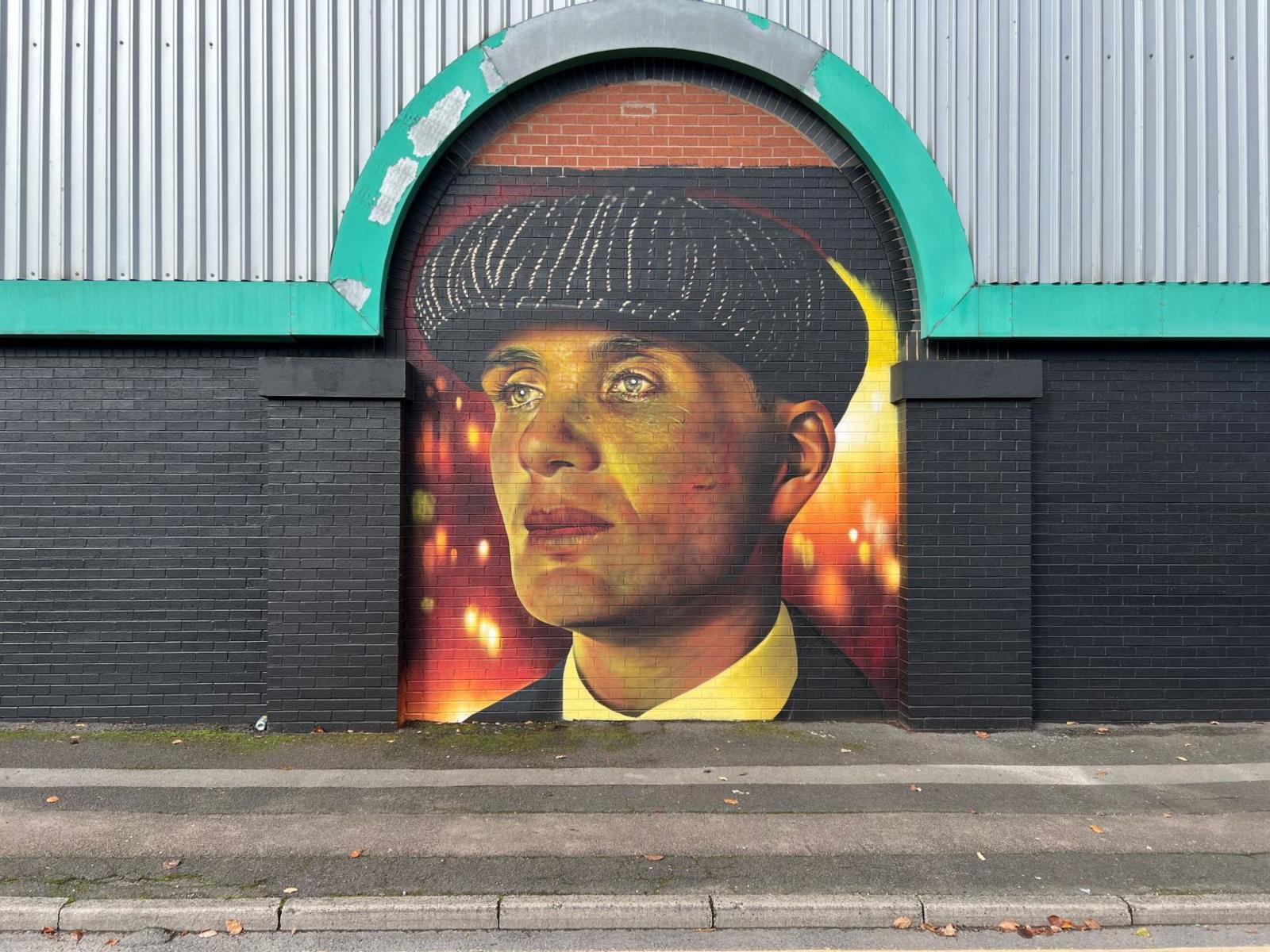 A street with a large colourful mural painted on a redbrick wall. The mural shows a close-up of Tommy Shelby from Peaky Blinders, a man wearing a black and white tweed flat cap, a black jacket and a white shirt. The mural uses yellow and orange tones on Shelby's face and has fire in the background.