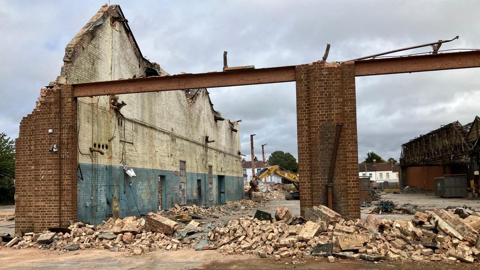 Crumbling side wall with bricks in piles near a former entrance. The bus depot yard can be seen with a digger in the distance. Piles of construction debris can be seen.