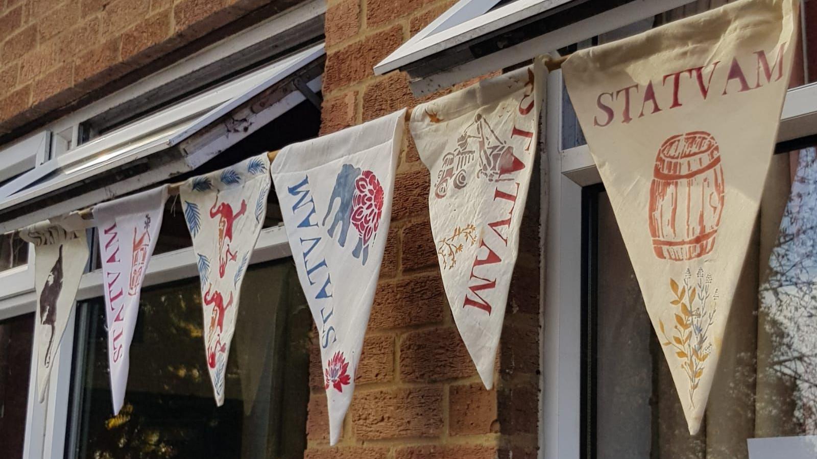 Bunting hanging below a window with various designs, including a barrel, an elephant and some flowers. Each triangle of bunting has the word STATVAM on it.