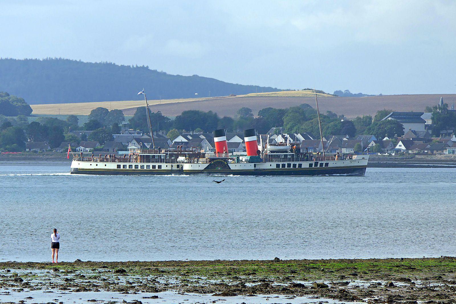 A shot of the red two-funnelled Waverly paddle steamer making its way along the River Tay. A woman is on the edge of the shore taking a picture.