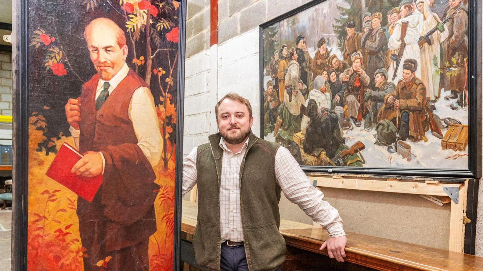 Joseph Trinder looks into the camera, wearing a check shirt and green gilet, while standing beside the tall painting of Lenin, which is quite a lot larger than him, to show its scale.