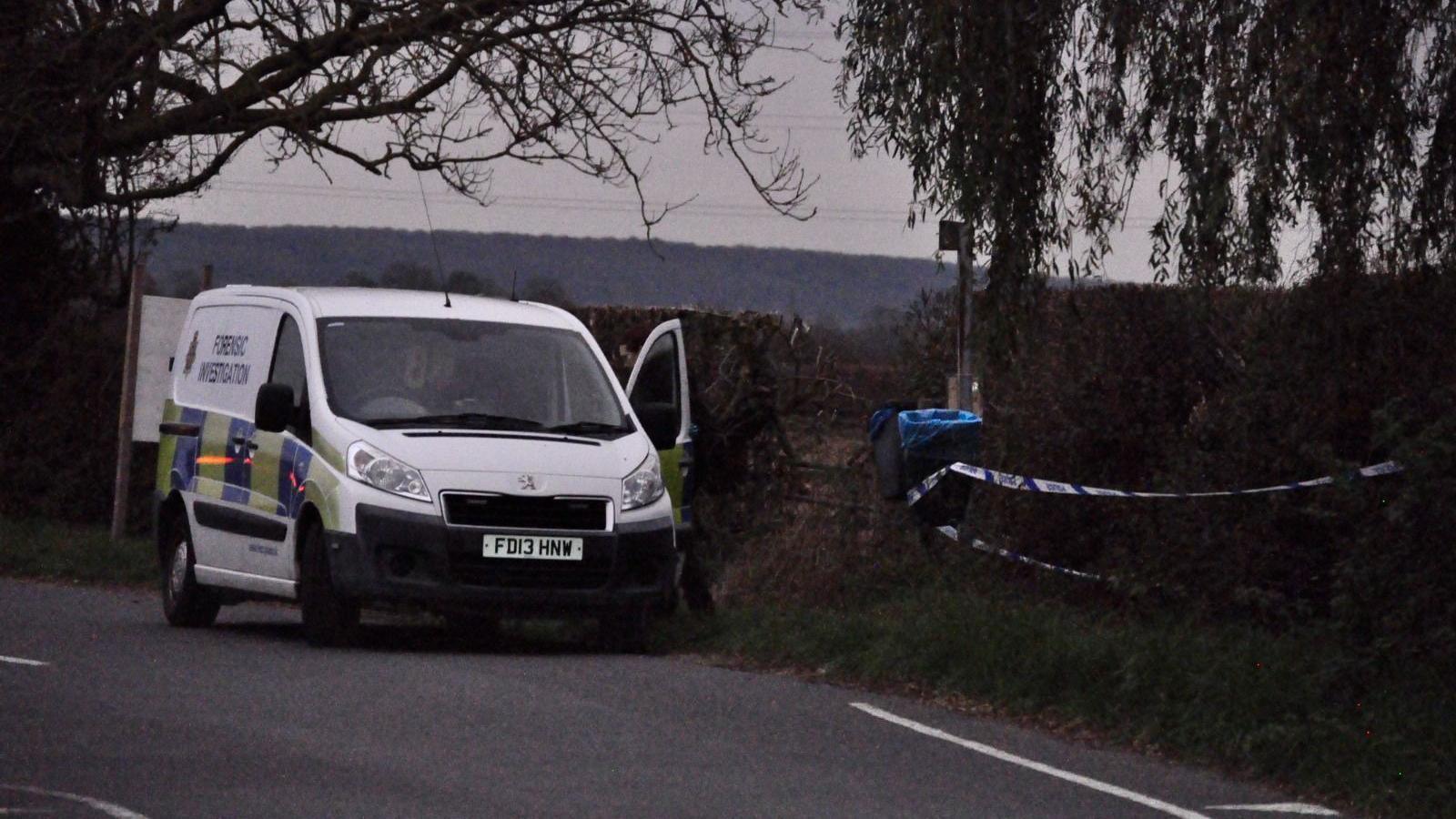 Police van, parked by a rural road, with trees and a high hedge, with police cordon tape visible along the hedge